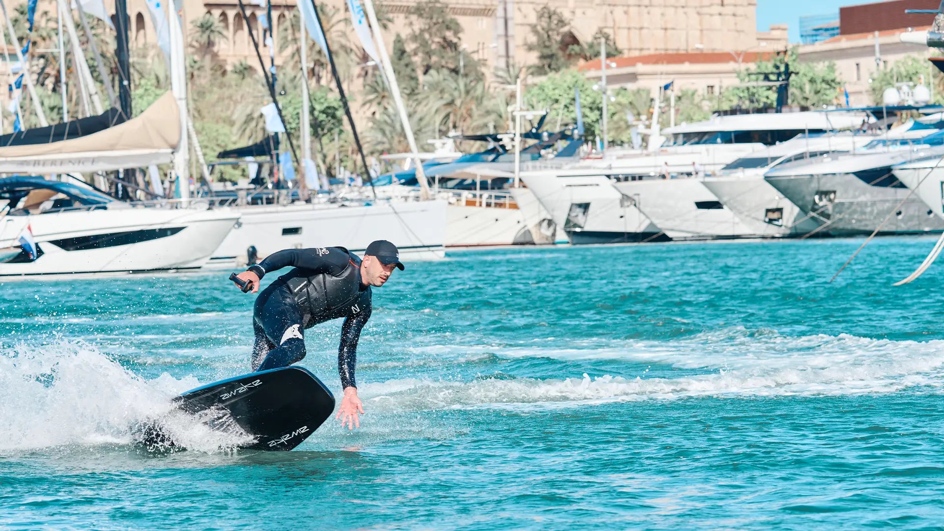 A man in a black neo rides an awake raevik ultimate elite on the water in the harbour of Palma de Mallorca, with many boats in the background.