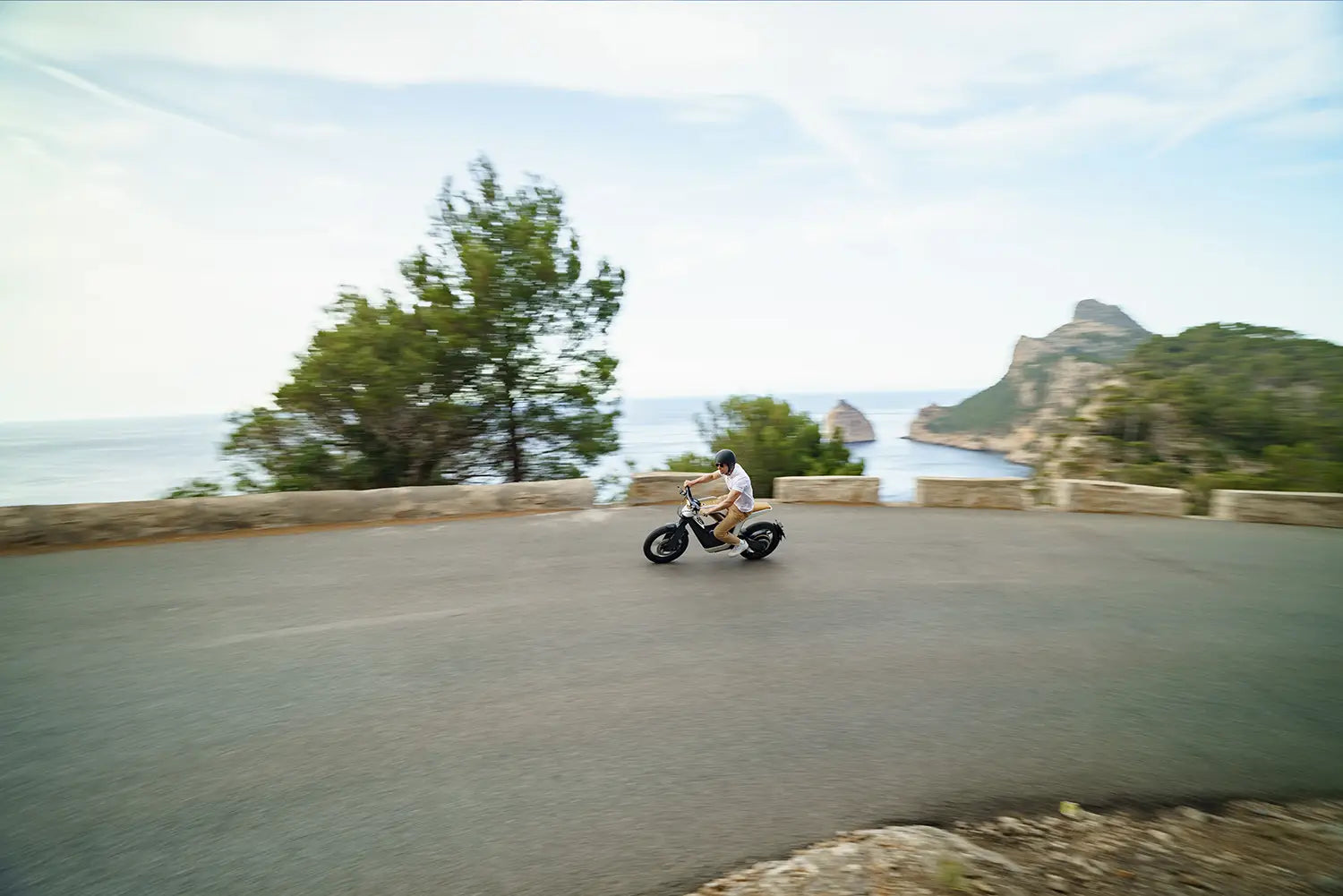 Person riding a NOVUS motorcycle on a scenic coastal road with trees and mountains in the background.