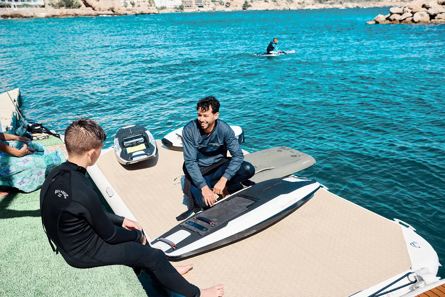 A surf instructor explains to a little boy how to ride an awake jetboard on the sea, in the background you can already see another person sitting on it.