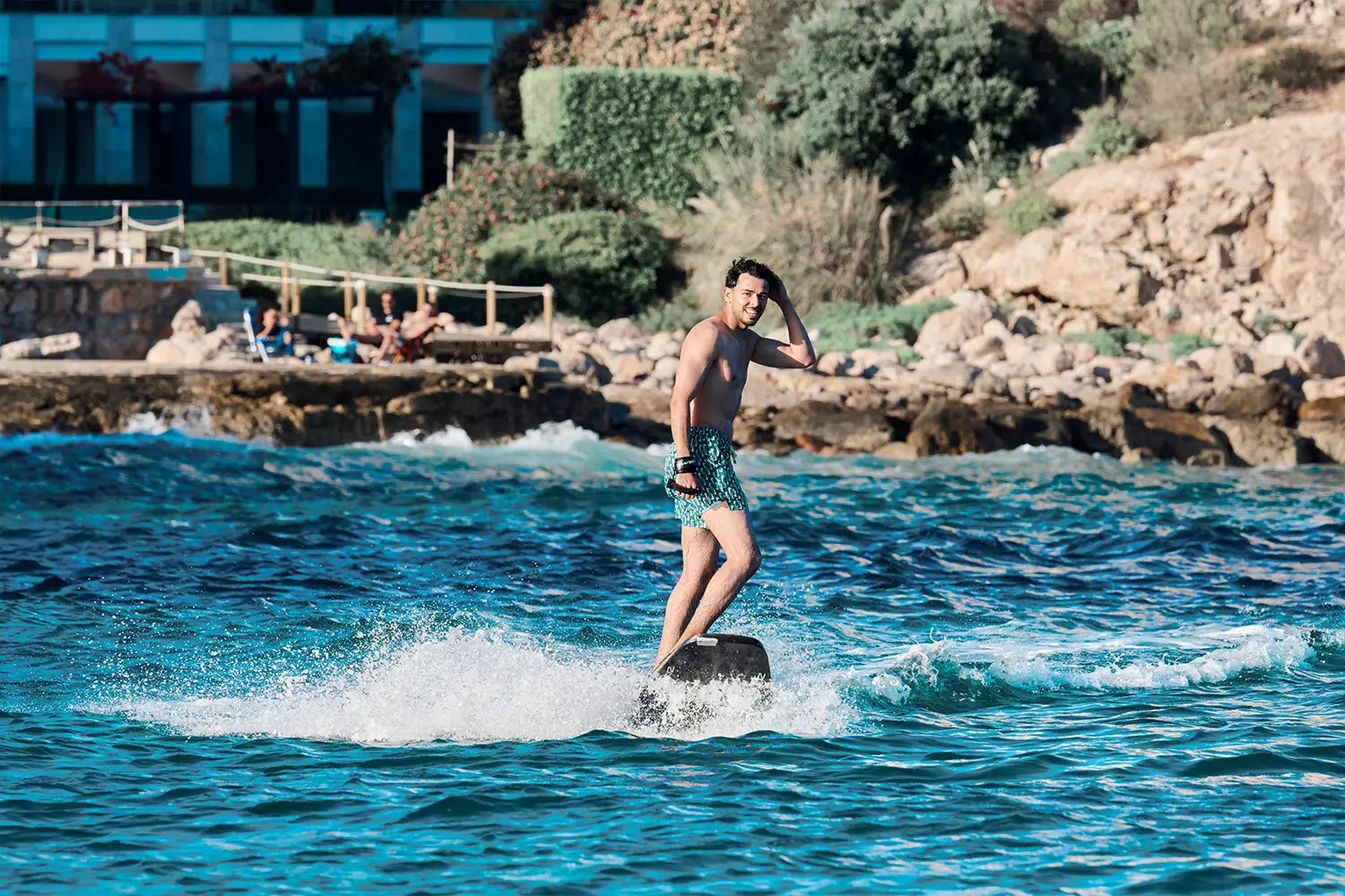 Ein Typ fährt mit einemjetboard die Kamerajetboard , im Hintergrund liegen Leute am Strand von Mallorca.