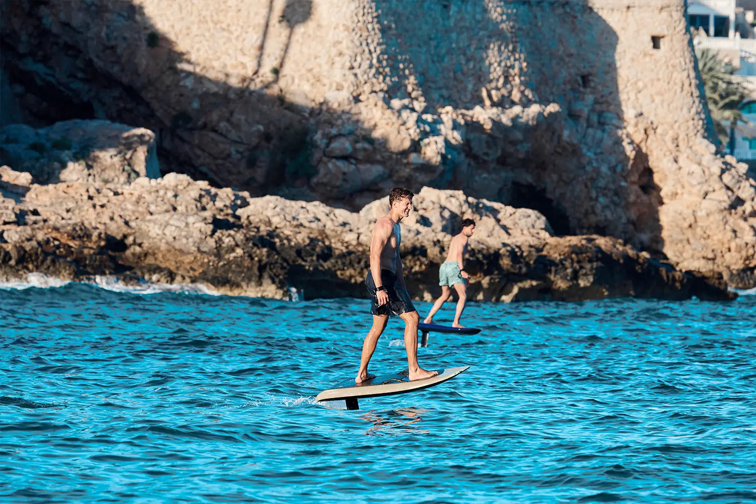 Two men fly over the water in Mallorca on a fliteboard, with brown rocks in the background.