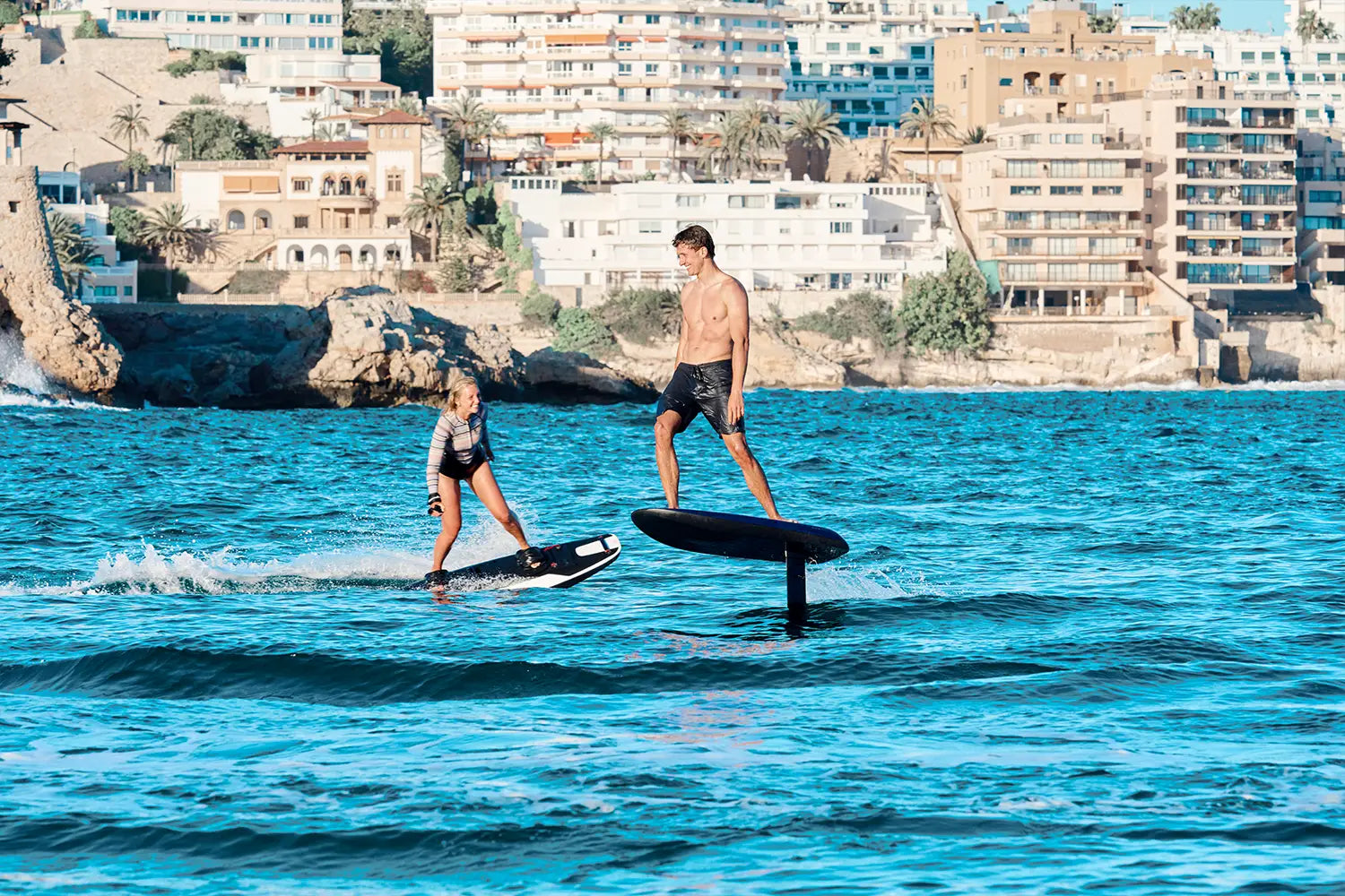 Ein Junge fährt fröhlich auf einem fliteboard auf dem Meer in Richtung einer Frau, die auf einemjetboard fährt, mit der Küstenlinie Mallorcas im Hintergrund.