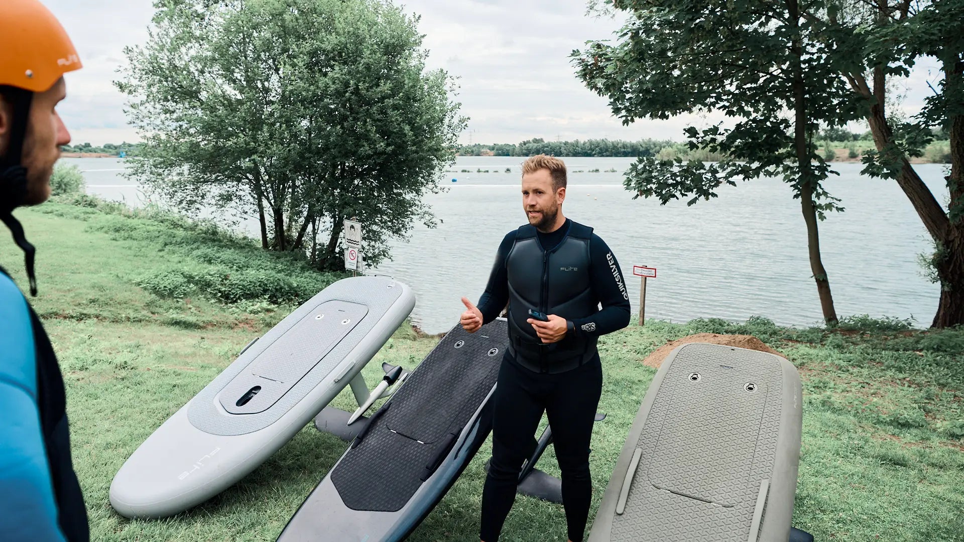 Man in a wetsuit explaining how to use a Fliteboard by a lake.