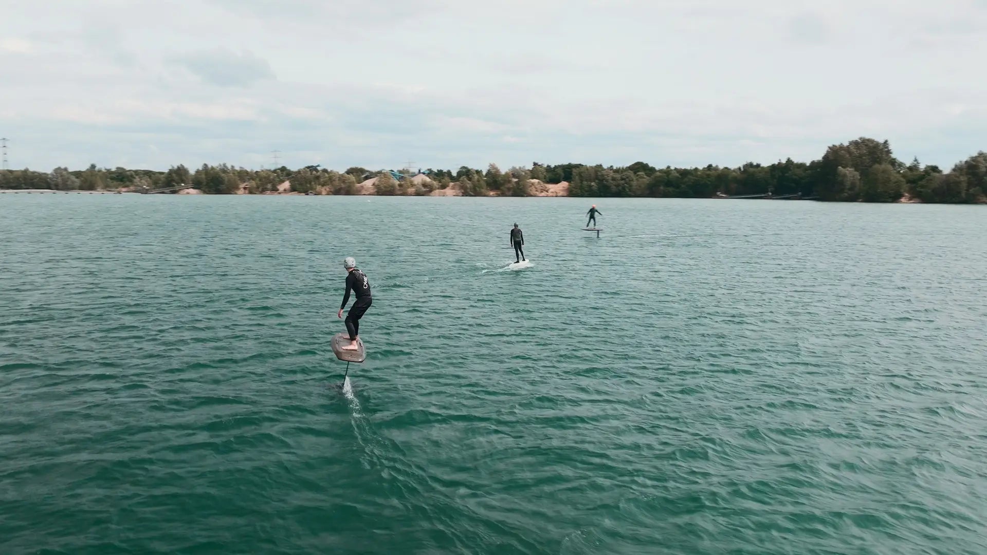 Three people efoiling on a calm lake with trees and buildings in the background.