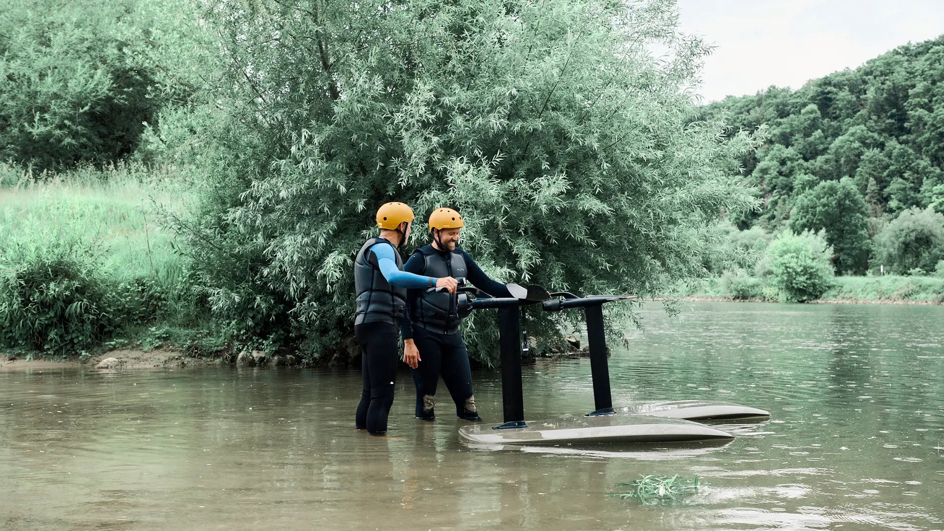 Two men stand up to their knees in the river, holding the wings of their fliteboards.