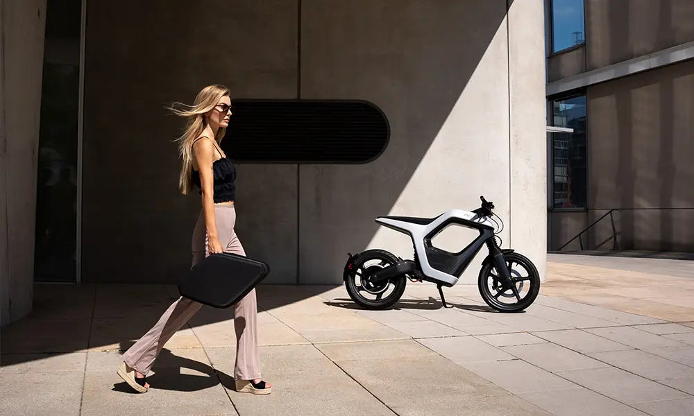 A woman walks down the street with a stylish bag, a white Novus bike in the background.