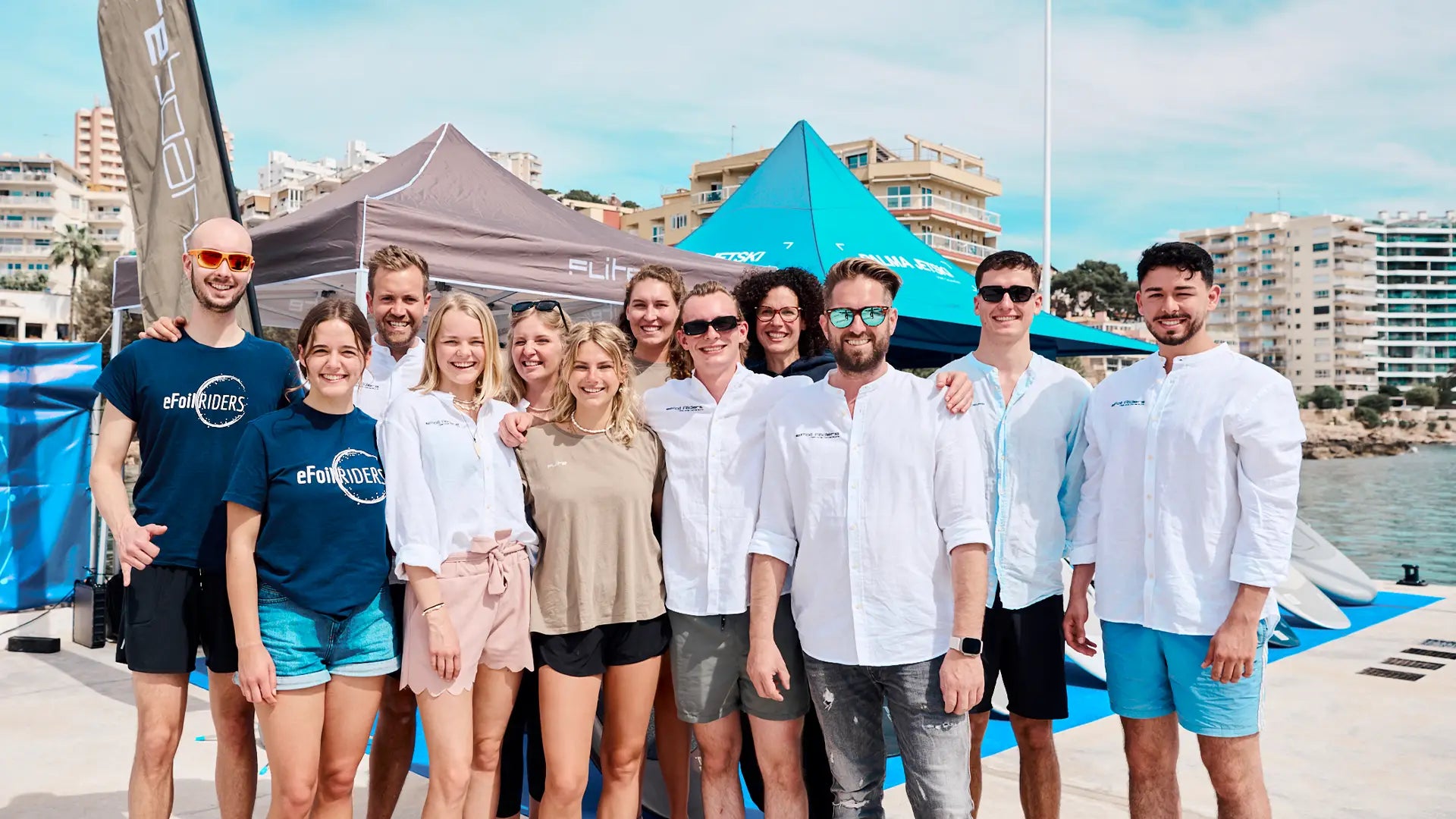 Group of people posing for a photo on a waterfront with tents and buildings in the background.