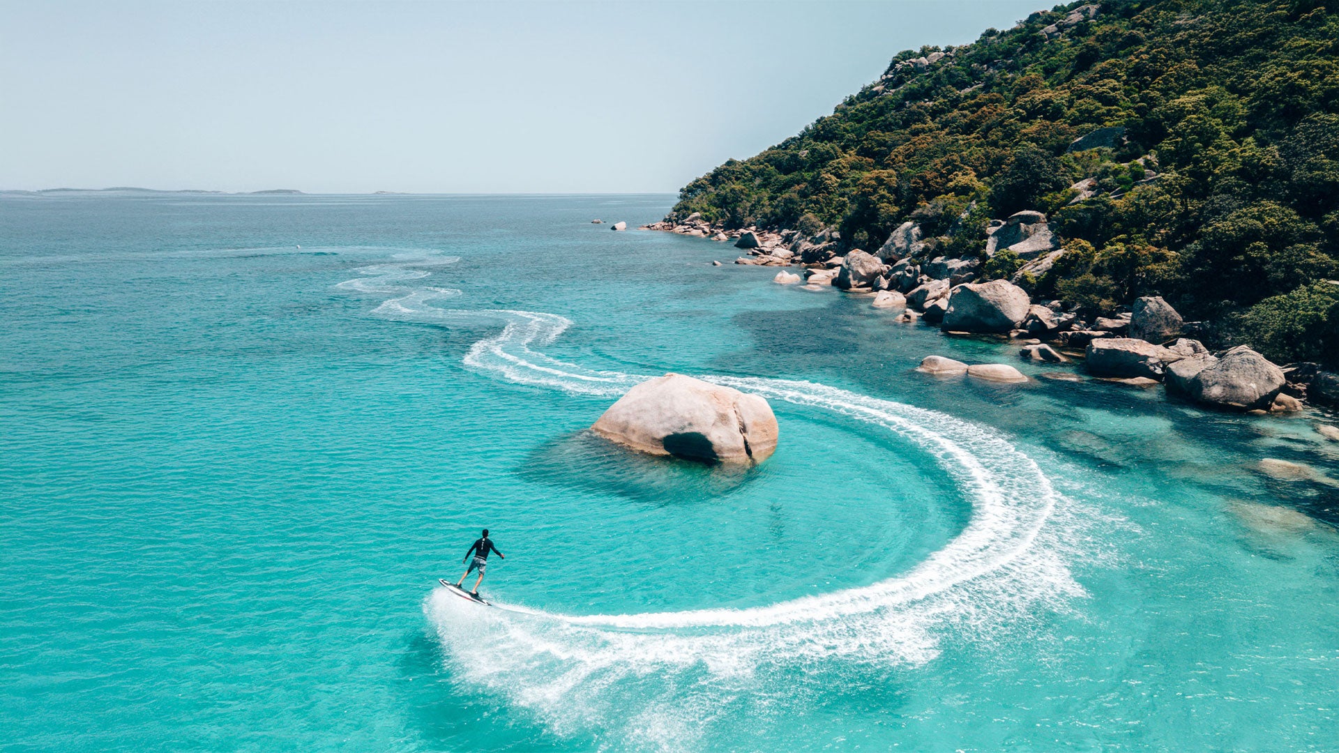 A bird's eye view of a person riding an awake raevik s jetboard round a rock on the sea.