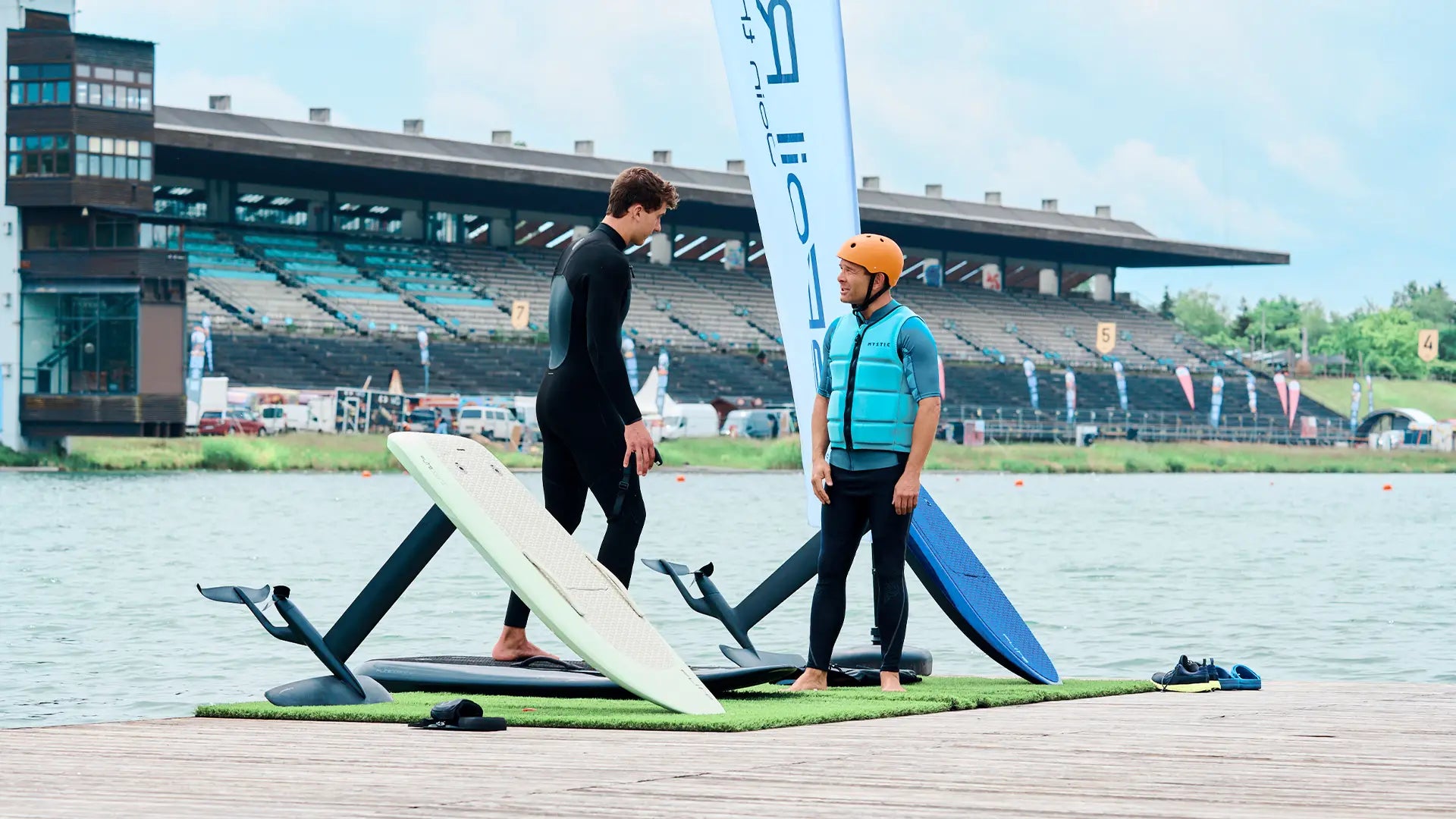 A surf instructor explains to his student on a pier how to ride a fliteboard, next to them are various boards and in the background you can see the regatta course in Munich.