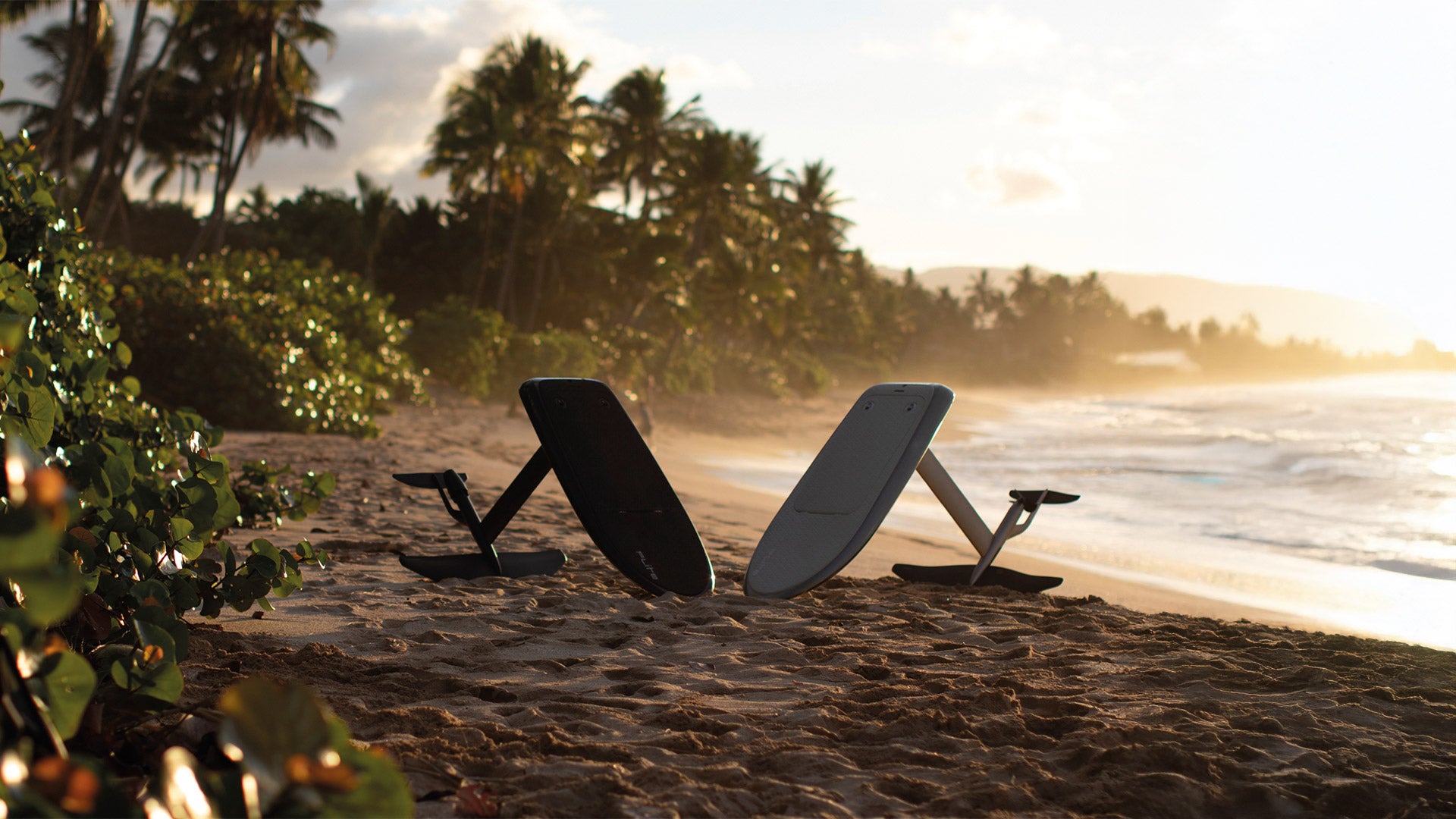 Two fliteboards, black on the left and white on the right, are set up on the beach while the sun sets in the background and transforms the tropical coastline into a beautiful light.
