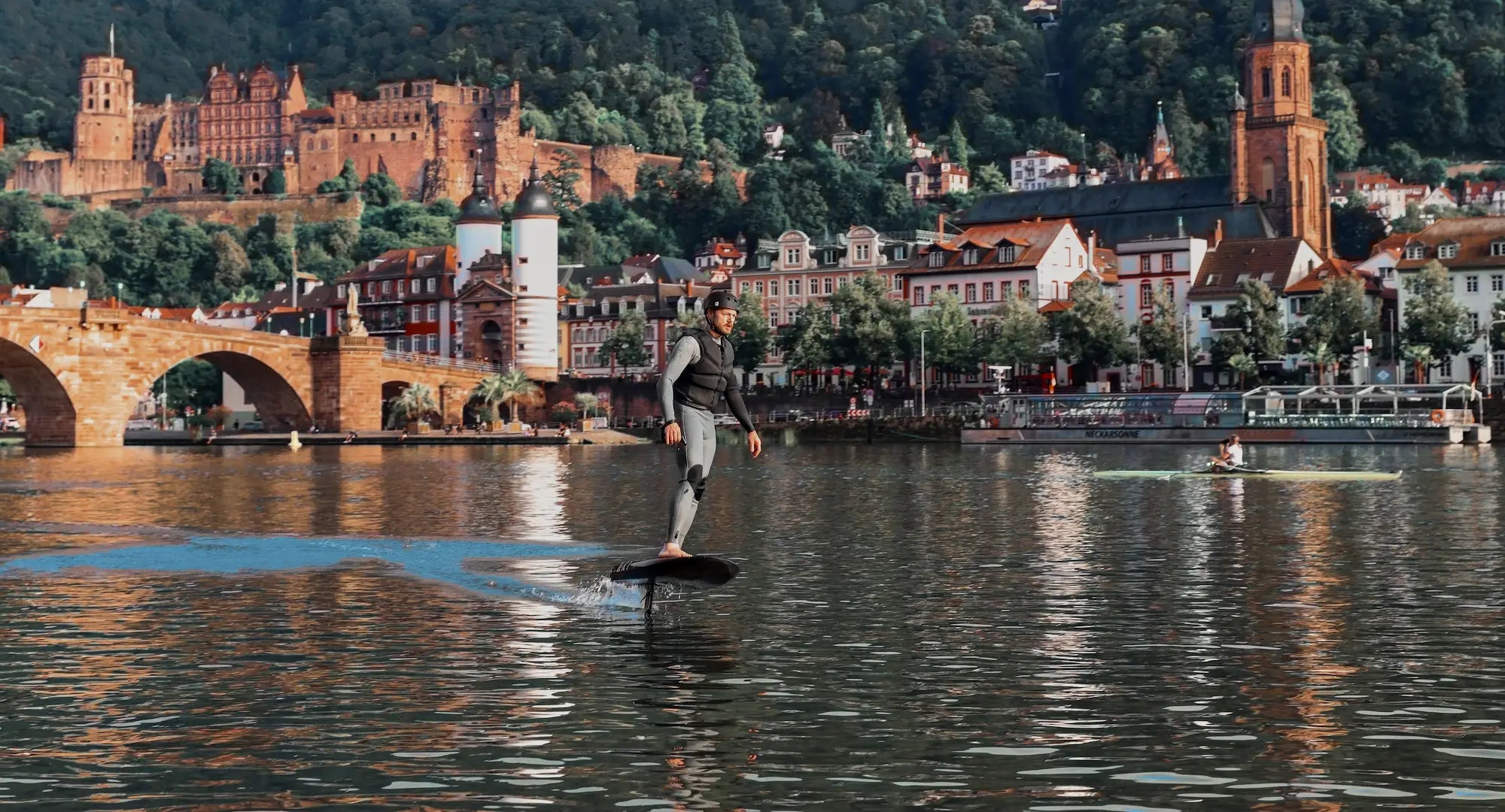 A man in a wetsuit and helmet flies over the river in Heidelberg on his fliteboard, with the castle and town in the background.