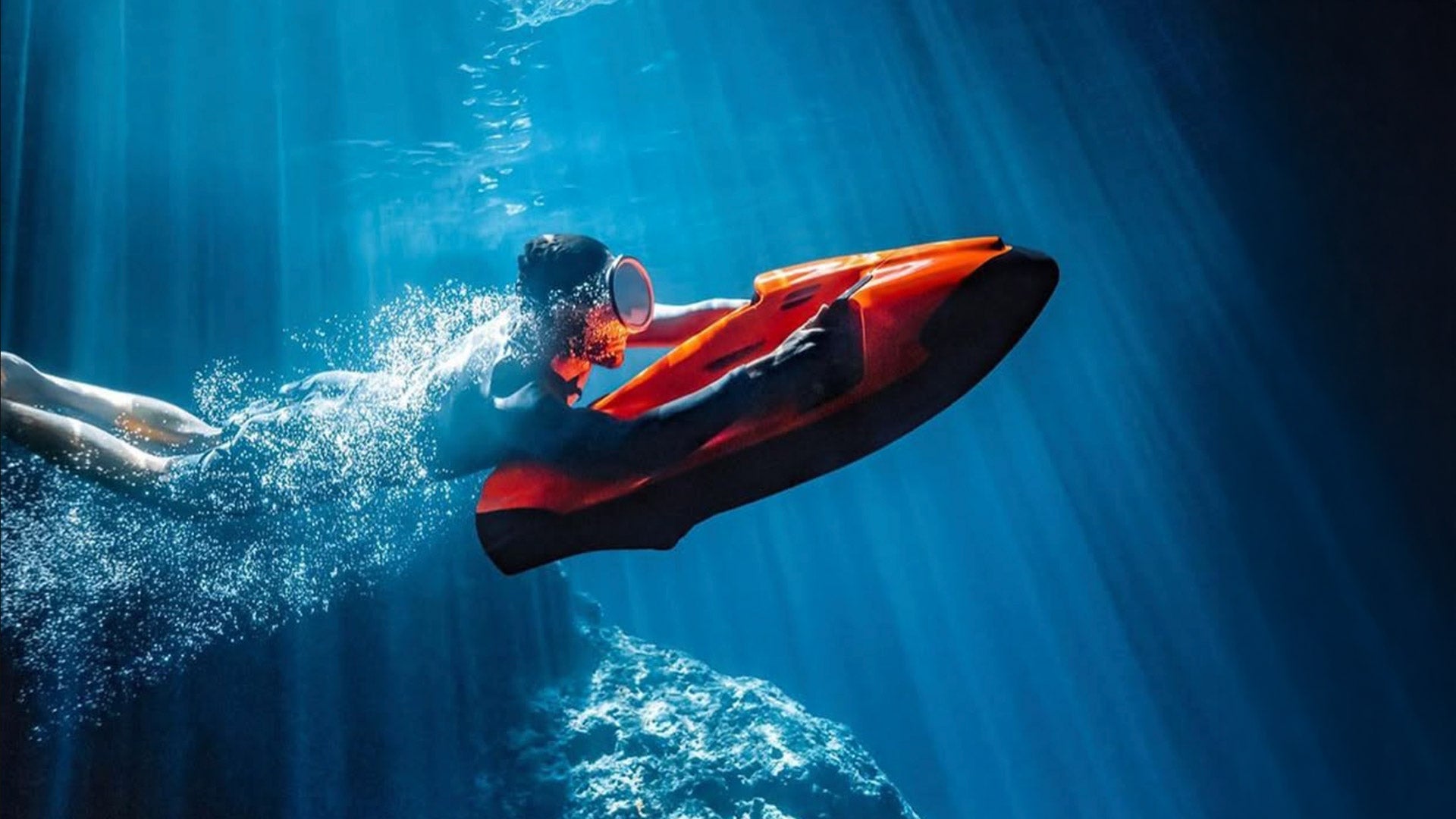 A person wearing diving goggles dives underwater with an orange Seabob.