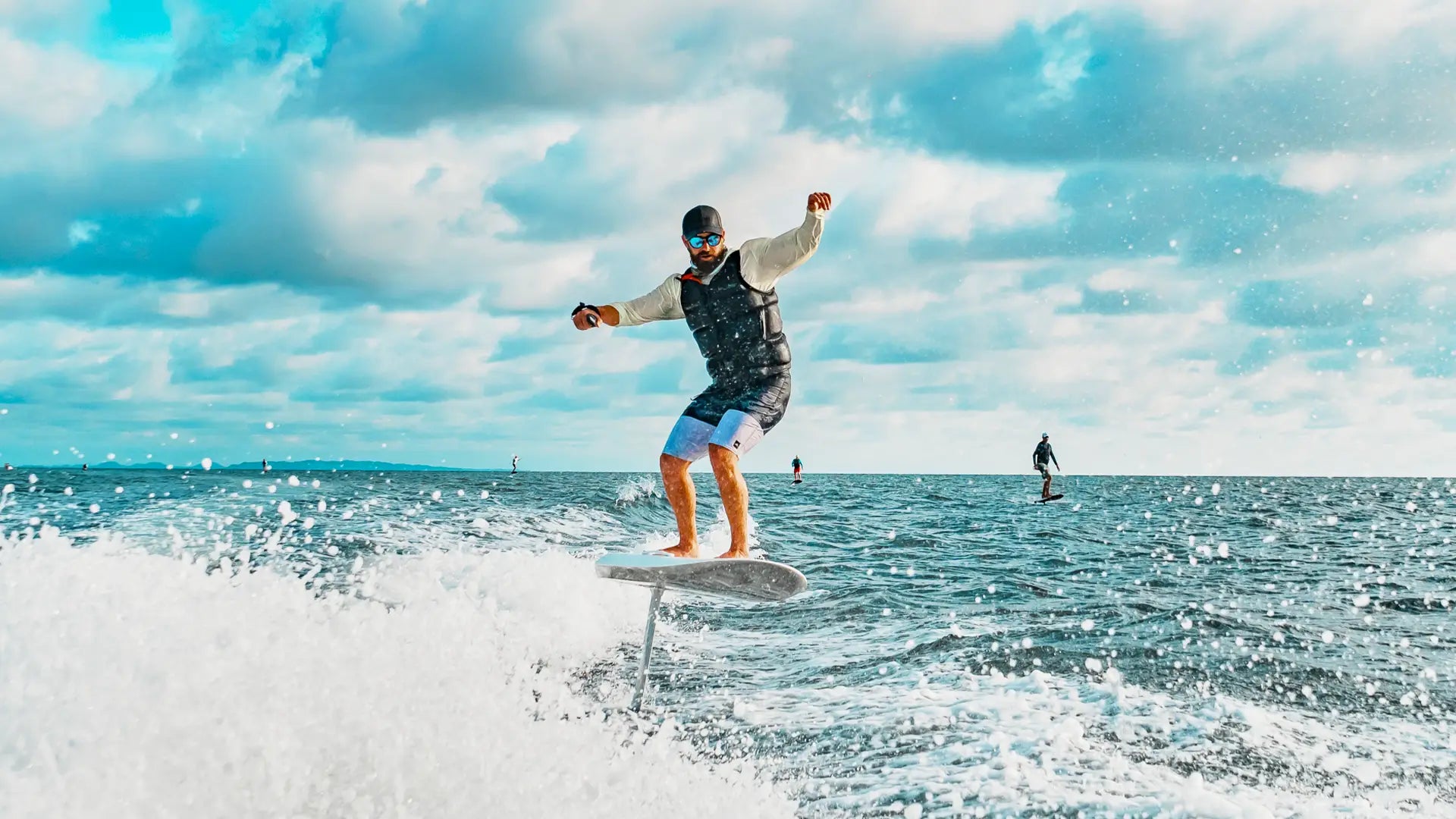 A man with sunglasses is efoiling on a fliteboard on the sea, in the background you can see other people efoiling.