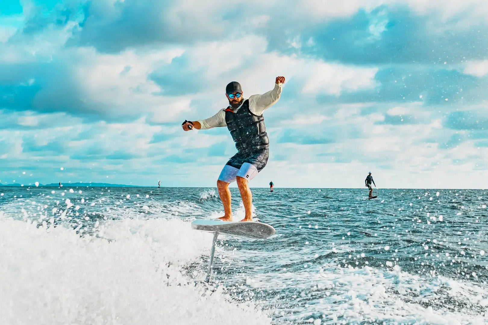 Ein Mann mit Sonnenbrille fährt mit einem Fliteboard auf dem Meer, im Hintergrund sind andere Leute zu sehen, die efoilen.