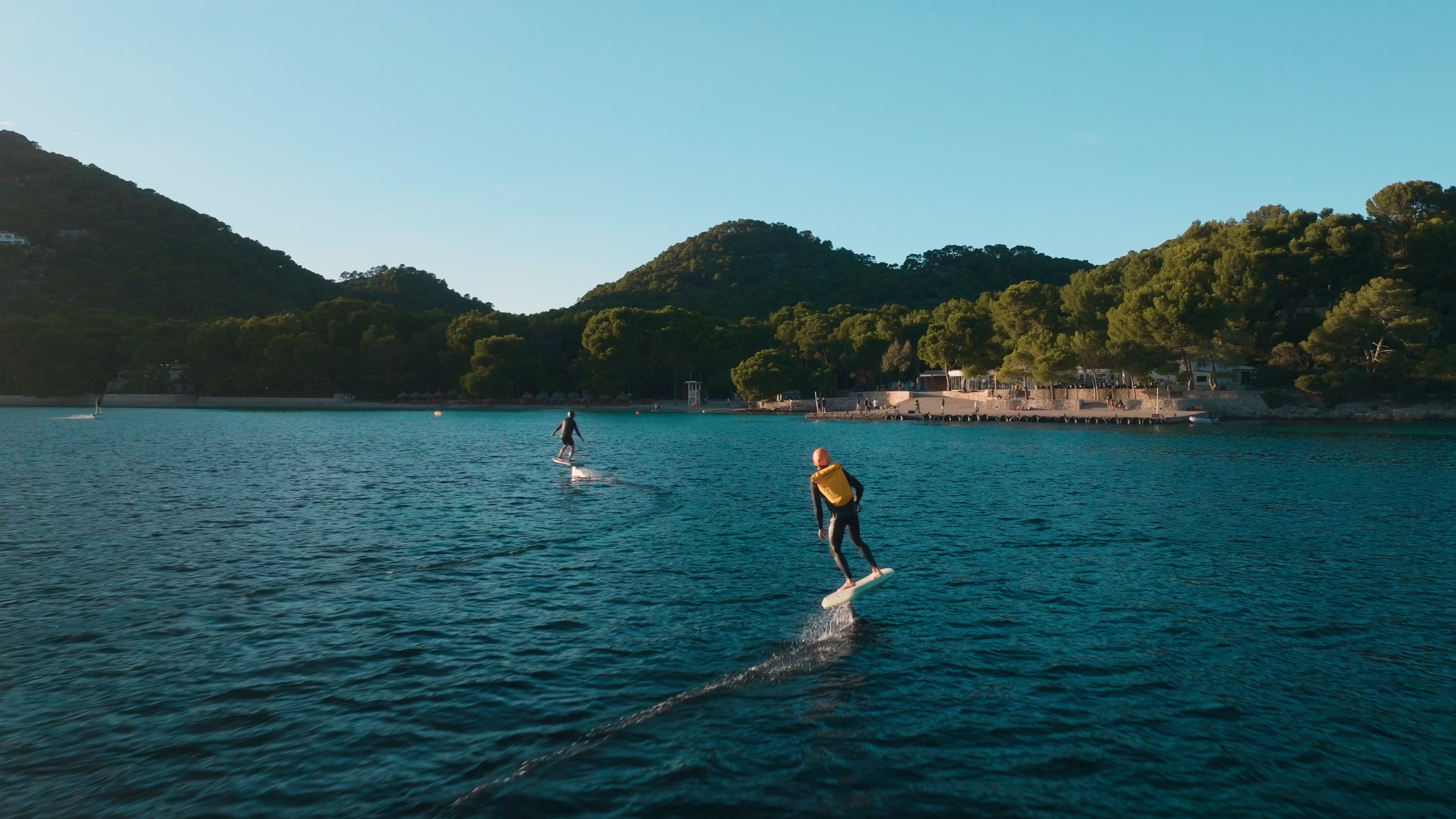 Two people are efoiling on the sea with fliteboards, in the background you can see mountains with green vegetation.