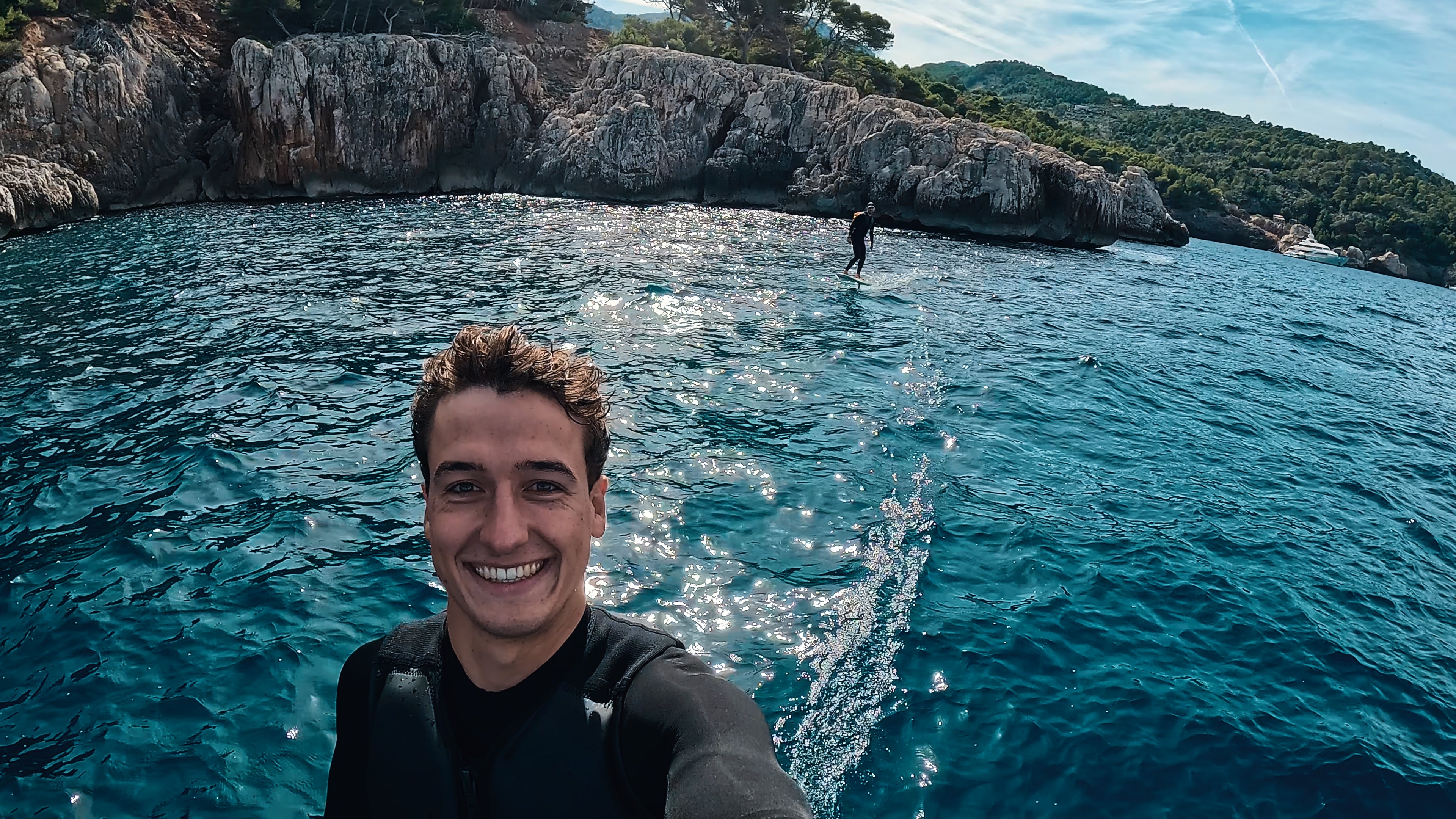 A person on the sea smiles into the camera while behind him another person is efoiling on a fliteboard.