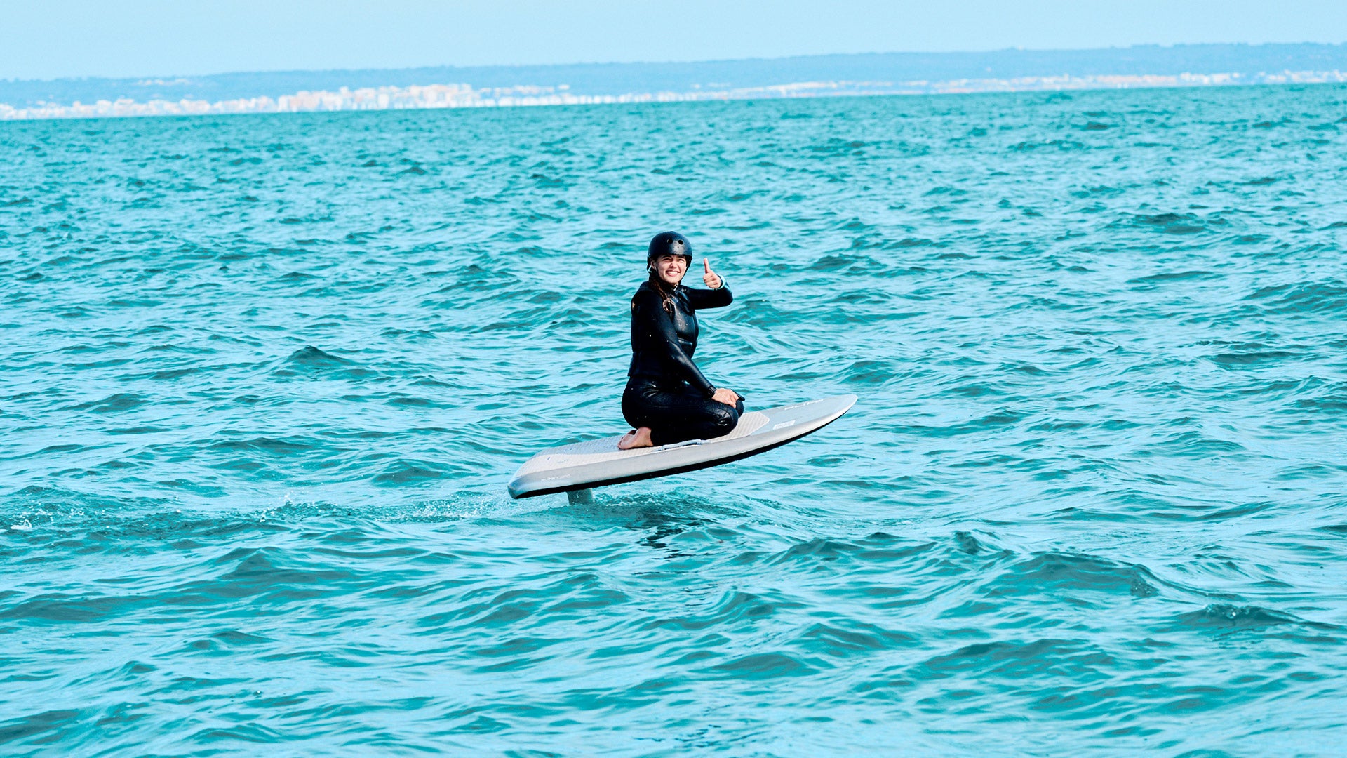A woman sits on a fliteboard and floats over the sea while pointing her thumbs up towards the camera.