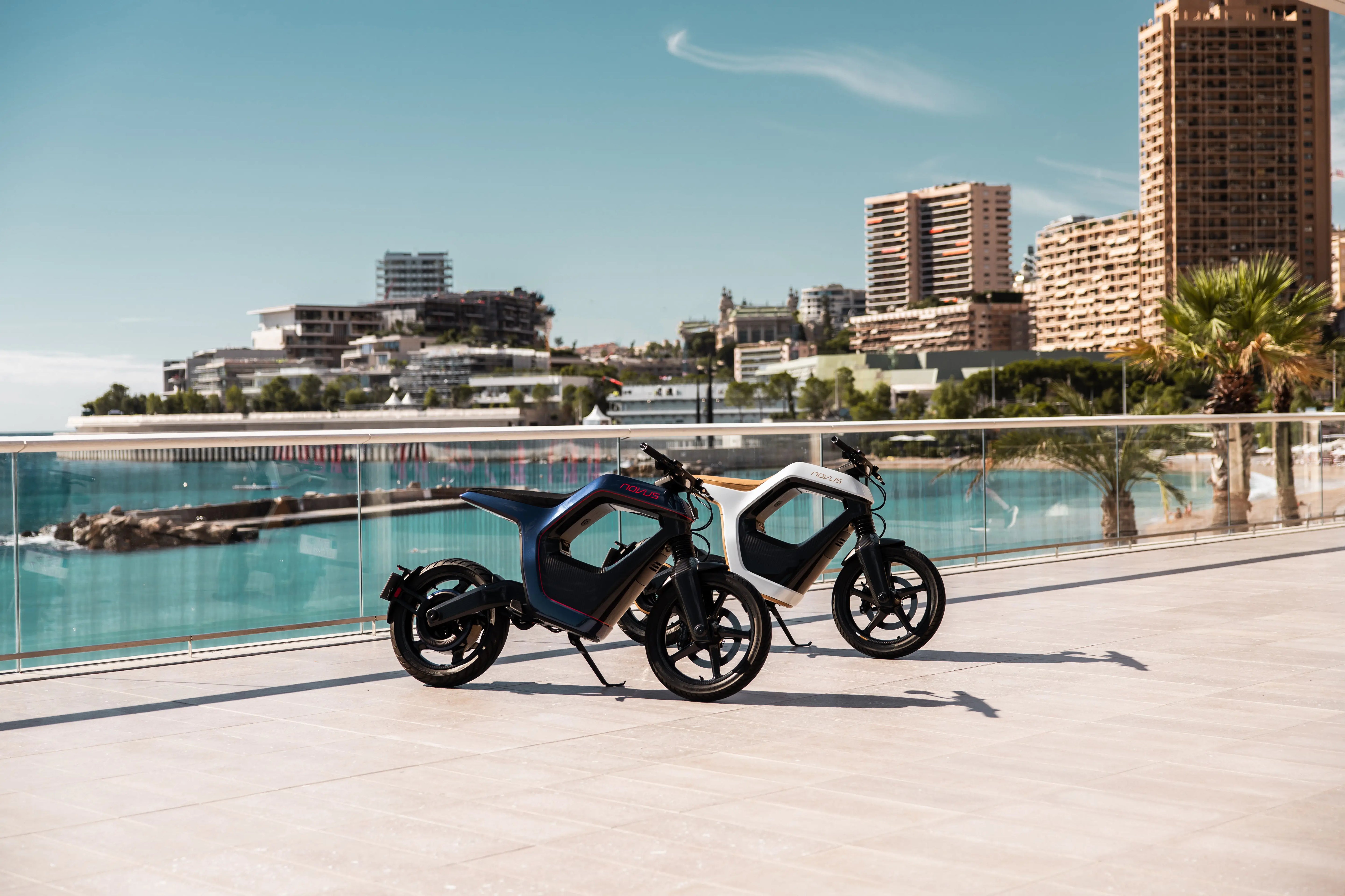 Two electric NOVUS motorcycles parked by a pool with a cityscape in the background.