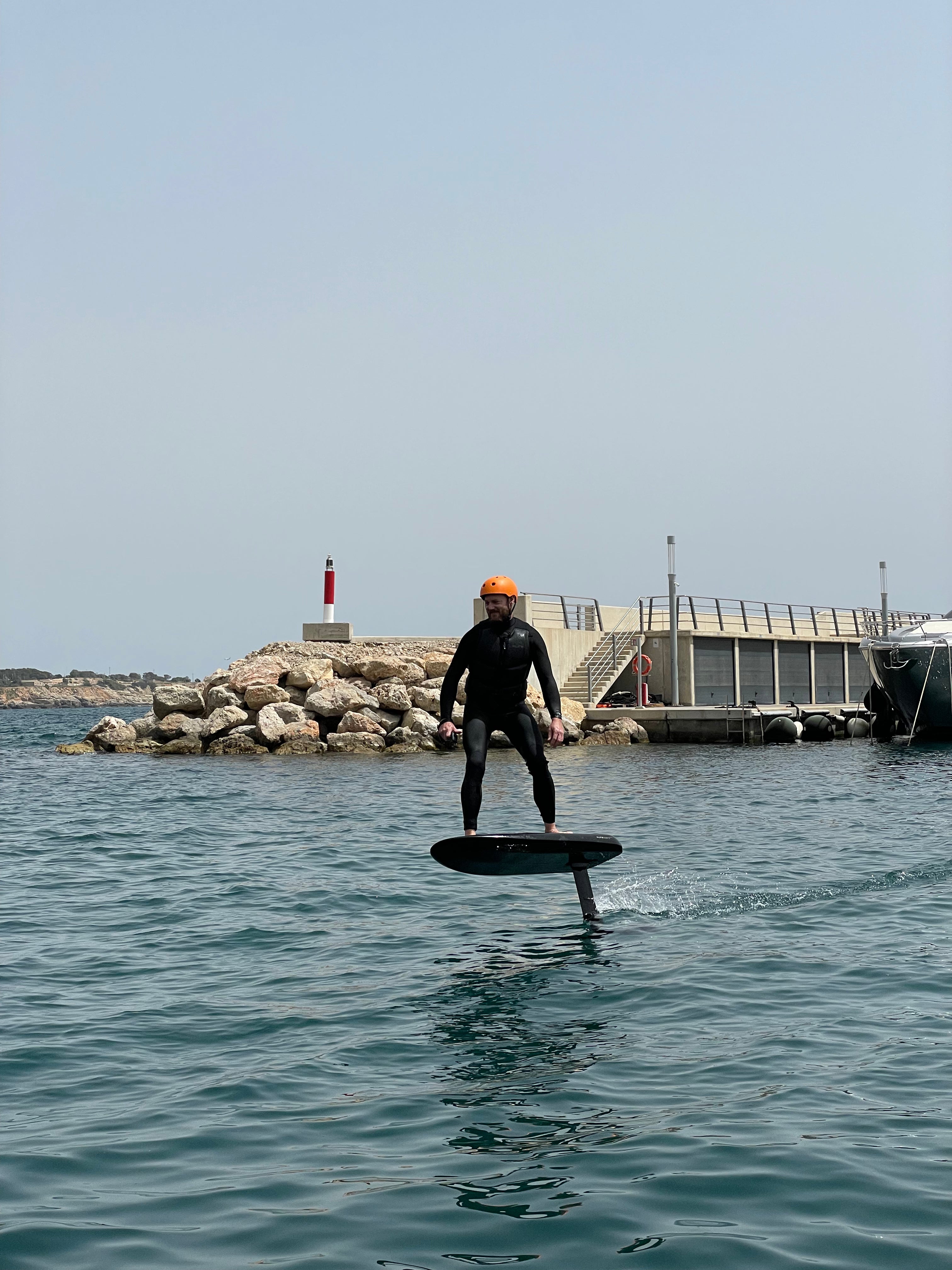 Jemand auf einem Fliteboard Wasser, mit einem Steg und einem Leuchtturm im Hintergrund.