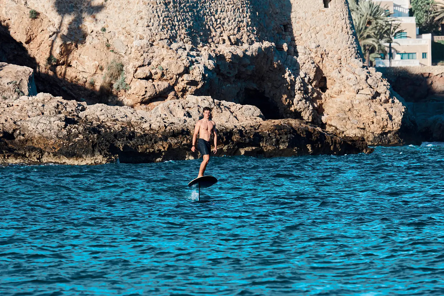 A boy efoils on a fliteboard on the sea in Mallorca, brown rocks can be seen in the background.