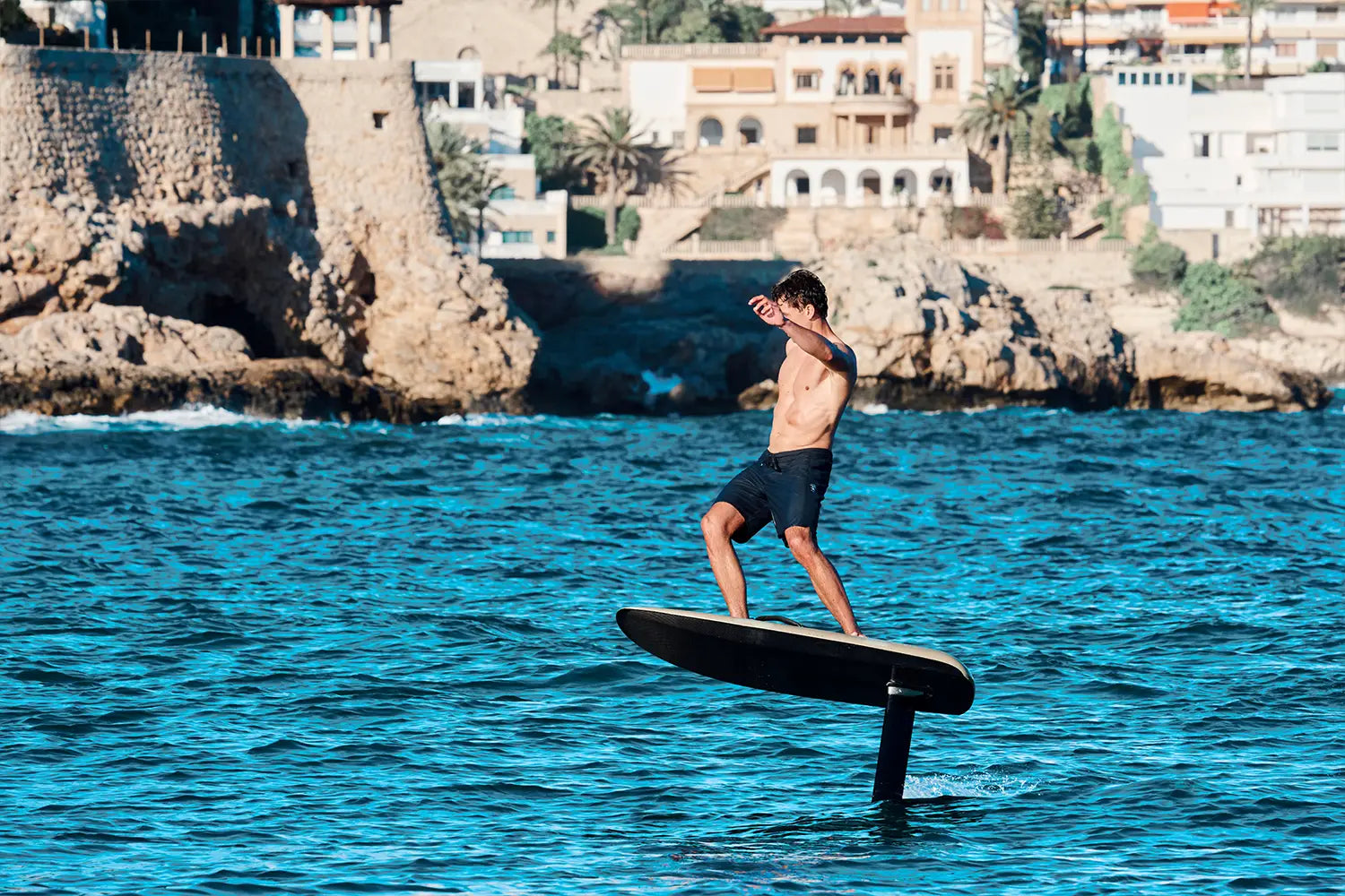 A boy efoils on a fliteboard on the sea in Mallorca, brown rocks can be seen in the background.
