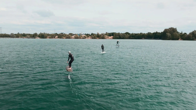 Drei Personen beim eFoilen auf einem ruhigen See mit Bäumen und Gebäuden im Hintergrund.