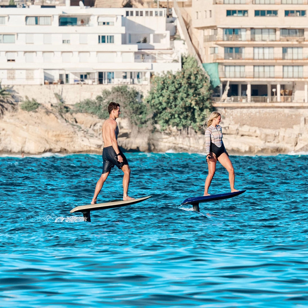 Two people on electric surfboards in the water with buildings in the background