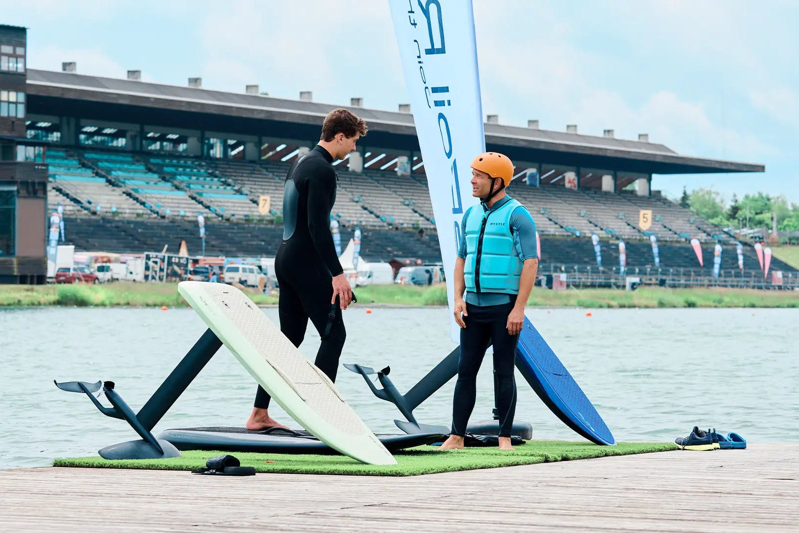Ein Surflehrer erklärt seinem Schüler auf einem Pier, wie man ein Fliteboard fährt. Neben ihnen stehen verschiedene Boards und im Hintergrund sieht man die Regattastrecke in München.