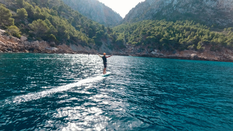 A person is efoiling on a fliteboard on the sea and holds his hands jubilantly towards the sky, in the background you can see another person efoiling and Mediterranean mountains.