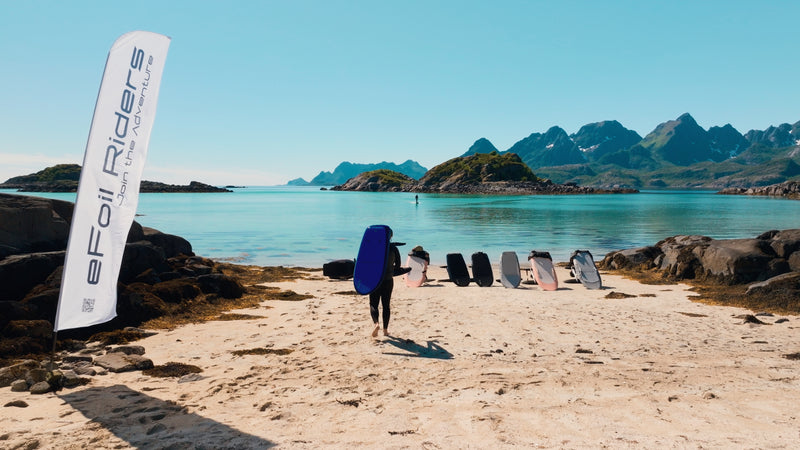 In the middle of Norway's Lofoten Islands, six fliteboards stand on the beach and a man carries the next one on his back, while a person is already efoiling in the background.